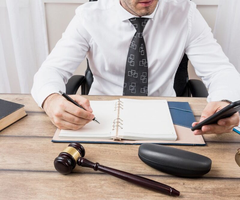 Pessoa assinando documento jurídico sobre mesa, ao lado de balança da justiça, representando o Provimento nº 206/2025 do CNJ que autoriza o acesso de juízes à CENSEC em casos de autocuratela.