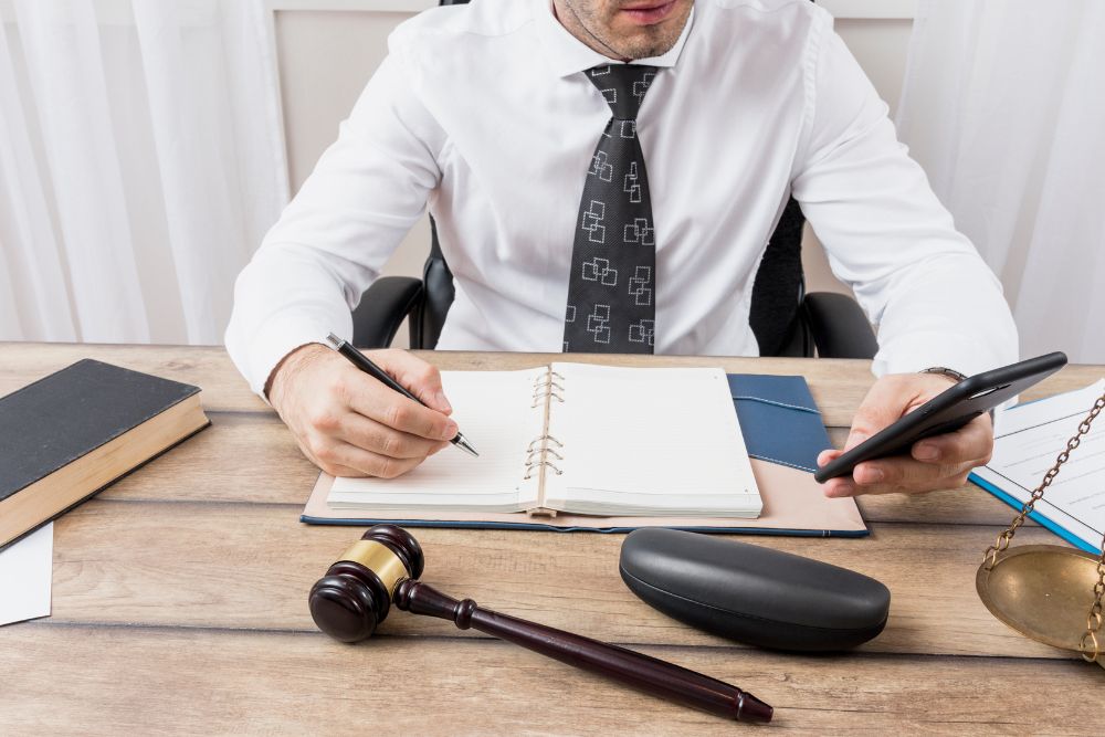 Pessoa assinando documento jurídico sobre mesa, ao lado de balança da justiça, representando o Provimento nº 206/2025 do CNJ que autoriza o acesso de juízes à CENSEC em casos de autocuratela.